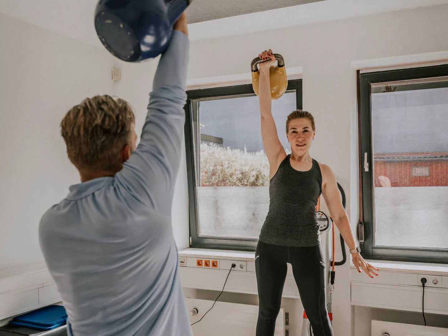 Eine Frau drückt eine Kettlebell über den Kopf, der Trainer macht die Bewegung ihr gegenüber mit